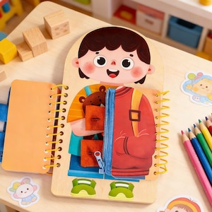 Puede incluir: Juguete educativo de madera con una ilustración de dibujos animados de un niño con una chaqueta roja y una mochila. El juguete tiene una cremallera, bolsillos y un pequeño oso de peluche. Las páginas están encuadernadas con espiral amarilla.