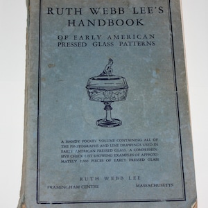 Op de afbeelding: Een blauw boek met de titel "Ruth Webb Lee's Handbook of Early American Pressed Glass Patterns". De omslag van het boek toont een zwart-witte illustratie van een glazen kom met een figuur erop.