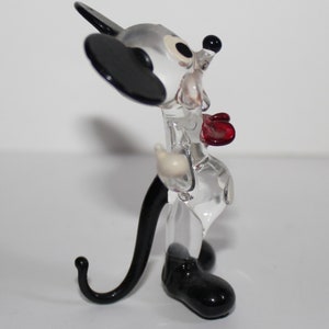 May include: A clear glass figurine of Mickey Mouse. The figurine is wearing a red bow tie and has black shoes and ears. The figurine is standing on a white surface.