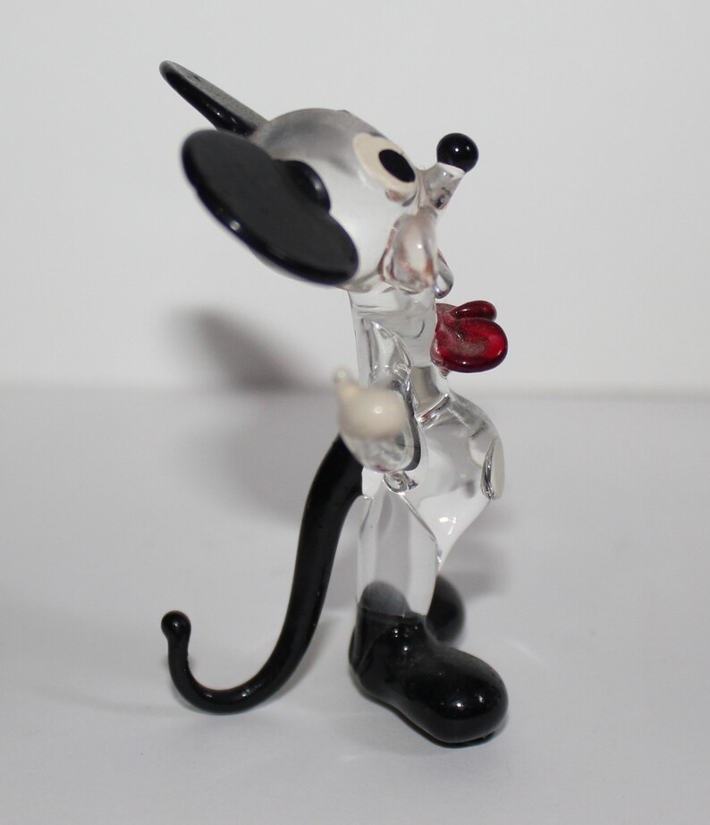 May include: A clear glass figurine of Mickey Mouse. The figurine is wearing a red bow tie and has black shoes and ears. The figurine is standing on a white surface.