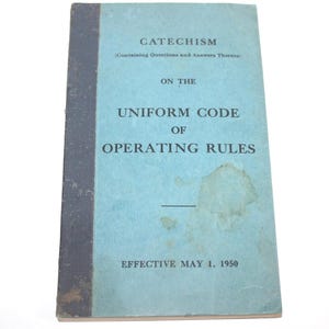 Op de afbeelding: Vintage boek getiteld "Catechismus over de Uniforme Code van Bedrijfsregels", met tekst in donkerblauw op een lichtblauwe kaft. De rug van het boek is donkerblauw. De publicatiedatum is 1 mei 1950.