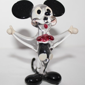 May include: A clear glass figurine of Mickey Mouse with black ears, shoes, and a bow tie. The figurine is in a standing pose with arms outstretched.