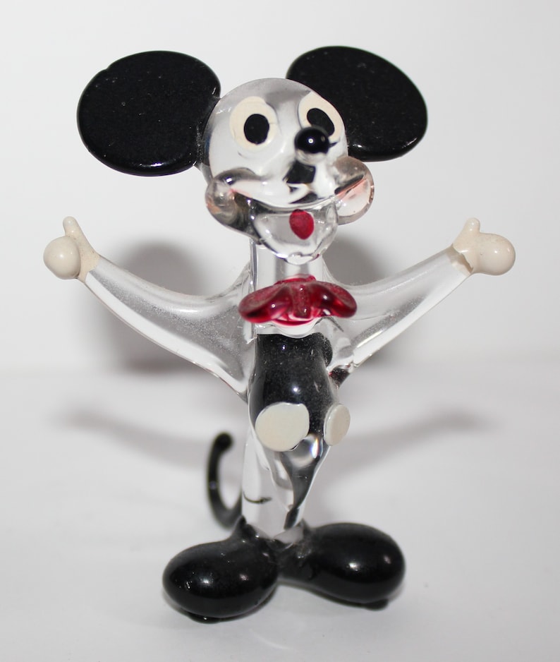 May include: A clear glass figurine of Mickey Mouse with black ears, shoes, and a bow tie. The figurine is in a standing pose with arms outstretched.