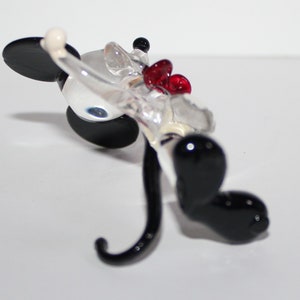 May include: A glass figurine of Mickey Mouse in a red bow tie. The figurine is clear glass with black painted ears, eyes, and shoes. The figurine is lying on its back with its tail curled up.