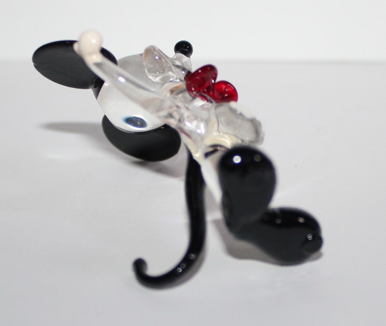 May include: A glass figurine of Mickey Mouse in a red bow tie. The figurine is clear glass with black painted ears, eyes, and shoes. The figurine is lying on its back with its tail curled up.