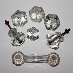 May include: A set of eight clear glass door knobs with a variety of styles, including round, square, and octagonal shapes. The knobs are all made of clear glass and have a polished finish. Some of the knobs have a metal screw attached to them.