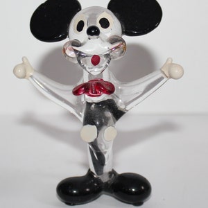 May include: A clear glass figurine of Mickey Mouse with black ears, red bow tie, and white accents. The figurine is standing with arms outstretched.