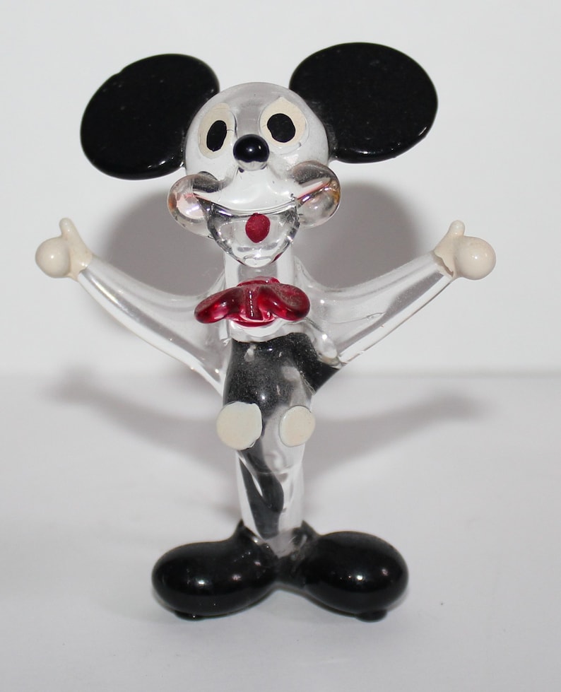 May include: A clear glass figurine of Mickey Mouse with black ears, red bow tie, and white accents. The figurine is standing with arms outstretched.