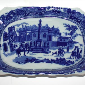 May include: A blue and white oval ceramic serving dish with a scalloped edge. The dish features a blue and white transferware design of a building with a column and figures in a landscape.