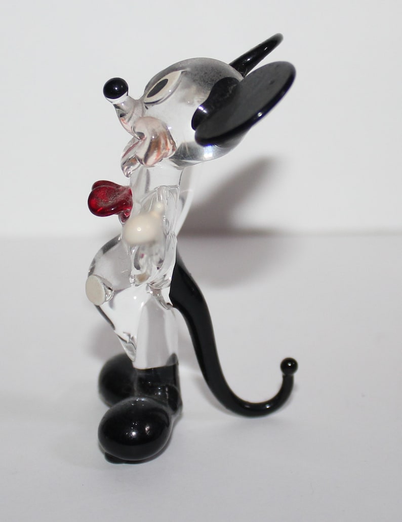 May include: A clear glass figurine of Mickey Mouse. The figurine is standing with its arms at its sides. Mickey is wearing a red bow tie and has black ears and shoes.