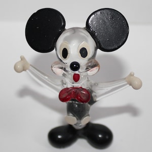 May include: A clear glass figurine of Mickey Mouse with black ears, red bow tie, and white gloves. The figurine is standing with its arms outstretched.