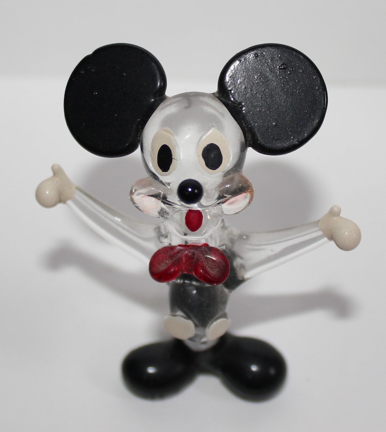 May include: A clear glass figurine of Mickey Mouse with black ears, red bow tie, and white gloves. The figurine is standing with its arms outstretched.