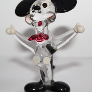 May include: A clear glass figurine of Mickey Mouse. The figurine is in a standing pose with arms outstretched. Mickey has black ears, black shoes, and white eyes. His mouth is open and he has a red tongue.