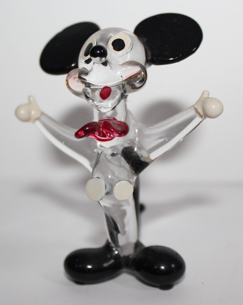 May include: A clear glass figurine of Mickey Mouse. The figurine is in a standing pose with arms outstretched. Mickey has black ears, black shoes, and white eyes. His mouth is open and he has a red tongue.