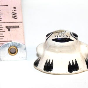 May include: A small, cream-coloured ceramic frog figurine with black painted details on its eyes, mouth, and feet. A pink measuring tape is positioned next to the figurine, showing a height of approximately 3.2 cm for the object.