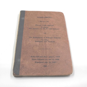 May include: A vintage brown booklet titled "AGREEMENT" between the Missouri Pacific Railroad and The Brotherhood of Railroad Trainmen. The booklet's cover has black binding and includes text about rules and rates effective in 1919 and 1966, respectively.