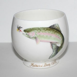 Könnte beinhalten: Weiße Keramiktasse mit einem handgemalten Bild einer Forelle, die mit einer Fliege im Maul aus dem Wasser springt. Die Tasse hat eine goldene Aufschrift auf dem Boden, die "Vatertag 1976" lautet.