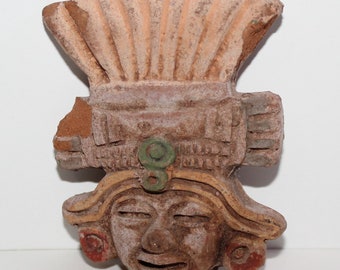 May include: A terracotta ceramic mask of a person with a headdress. The mask has green, yellow, and red paint details. The person has a wide mouth and closed eyes.