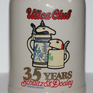Peut inclure: Une chope à bière en céramique blanche avec un motif rouge et or. La chope représente deux chopes à bière en dessin animé, l'une avec un motif floral bleu et blanc et l'autre avec un trèfle vert. Le texte "Utica Club", "35 Years", "Schultz & Dooley", et "Limited Edition" est imprimé sur la chope.