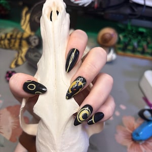 Może przedstawiać: Dłoń z długimi, czarnymi paznokciami w kształcie migdałów, ozdobionymi żółtymi i złotymi wzorami, w tym półksiężycem i kotem. Dłoń trzyma białą czaszkę z kości.