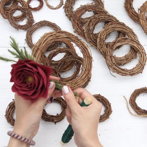 May include: A collection of small, brown grapevine wreaths in various sizes, some intertwined. A person is holding a red rose with green foliage, preparing to attach it to a wreath. A ball of twine is also visible.