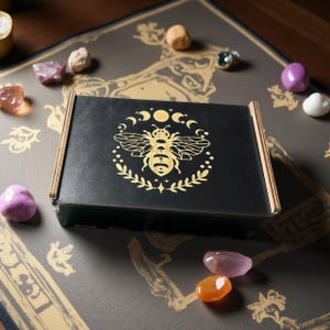 Peut inclure: Une boîte noire avec un motif d'abeille dorée, entourée de lunes et d'étoiles, est posée sur une surface à motifs. Diverses pierres précieuses colorées sont éparpillées autour de la boîte. La boîte a un cadre en bois.
