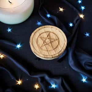 May include: A round, wooden coaster with a carved pentagram design. The coaster is light brown and sits on a dark blue fabric with star-shaped lights. A lit, white candle is in the upper left corner.