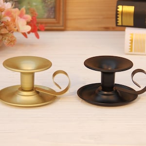 Może przedstawiać: Dwa metalowe świeczniki, jeden złoty i jeden czarny, stoją obok siebie. Każdy ma okrągłą podstawę, centralną nóżkę i mały uchwyt. Złoty uchwyt ma błyszczące wykończenie, a czarny matowe. Przedmioty dekoracyjne.