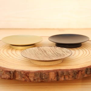 May include: Three small, round candle holders in gold, black, and a textured bronze finish. The candle holders are displayed on a natural wood slice with a light brown background.