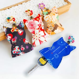 Può includere: Quattro coprichiavi in tessuto a forma di sagome di gatto, ognuna con un motivo colorato unico. Le coperture includono stampe floreali nere, rosse e beige, e una stampa blu con fuochi d'artificio, tutte sormontate da piccoli fiori in tessuto.