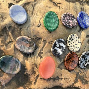 Puede incluir: Una variedad de piedras preciosas pulidas en varios colores y patrones, incluyendo azul, verde, rosa y negro, dispuestas sobre una superficie texturizada de color marrón claro. Las piedras tienen forma ovalada y varían en tamaño y textura.