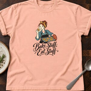 May include: A peach-colored t-shirt featuring a graphic of a person with a yellow headband wearing a blue shirt and black apron, holding a baking sheet of cookies and eating one. The text "Bake Stuff, Eat Stuff" is printed in black script below.