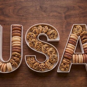 May include: The word "USA" is spelled out with white serving dishes filled with cookies and macarons. The "U" and "A" are filled with macarons, while the "S" is filled with chocolate chip cookies. The background is a dark wood surface.