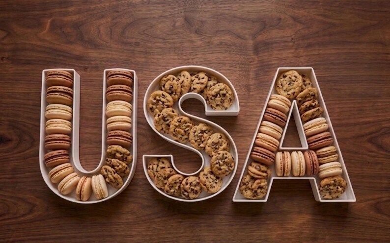 May include: The word "USA" is spelled out with white serving dishes filled with cookies and macarons. The "U" and "A" are filled with macarons, while the "S" is filled with chocolate chip cookies. The background is a dark wood surface.