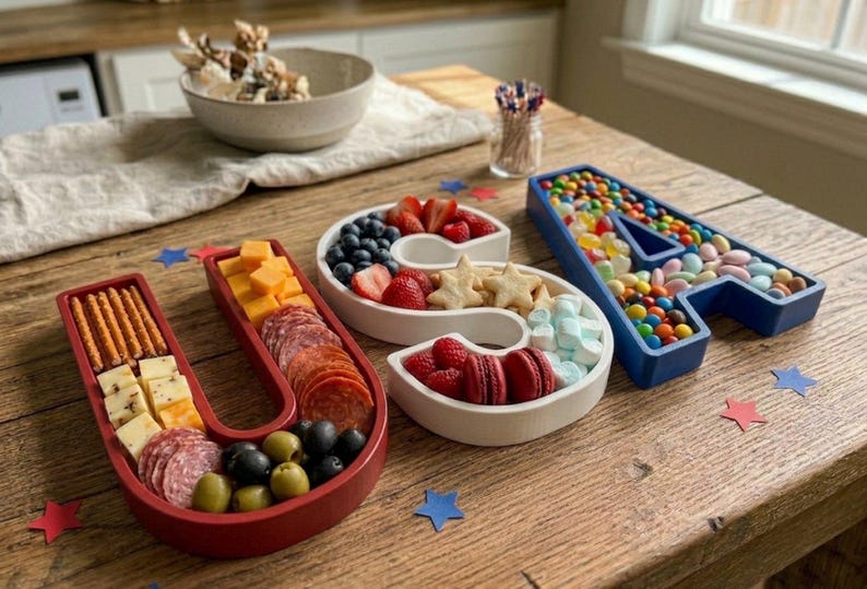 May include: A wooden table displays a patriotic-themed food arrangement spelling "USA". The red "U" holds crackers, cheese, salami, and olives. The white "S" contains berries, cookies, and macarons. The blue "A" is filled with colorful candies.