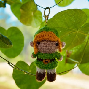 Può includere: Un piccolo portachiavi a forma di bambola amigurumi all'uncinetto che raffigura un personaggio con capelli giallo oro, una tunica verde, una cintura marrone scuro con fibbia gialla, pantaloni bianchi e stivali marroni, con un moschettone dorato.