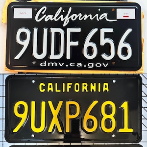May include: Two black California license plates. The top plate has white lettering and numbers, with the text "California" in cursive. The bottom plate has yellow lettering and numbers. Both plates display the DMV website.