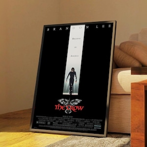 May include: Framed movie poster for "The Crow" featuring Brandon Lee. The poster has a black background with the words "Believe in Angels" and an image of a person walking towards the viewer. The title "the crow" is in red.