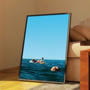 May include: Framed art print depicting a serene ocean scene under a clear blue sky. Two figures relax on a yellow inflatable raft in the water. A large, donut-shaped object floats nearby. The artwork is displayed on a wall.