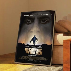 May include: A framed movie poster for "No Country for Old Men" featuring a large face in the background and a running figure in the foreground. The poster has the film's title and the names of the actors.