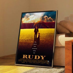 May include: Framed movie poster for the film "Rudy." The poster features a man in a suit standing on a football field, with a stadium in the background. The title "RUDY" is prominently displayed at the bottom, with text that reads "When people say dreams don't come true, tell them about."