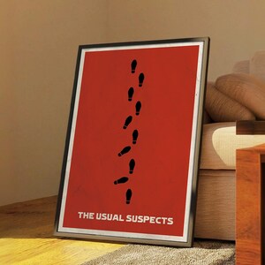 Op de afbeelding: Ingelijste poster met een rode achtergrond met een reeks zwarte voetstappen die verticaal oplopen. De tekst "THE USUAL SUSPECTS" is in het wit onderaan gedrukt. De poster zit in een zwart frame.