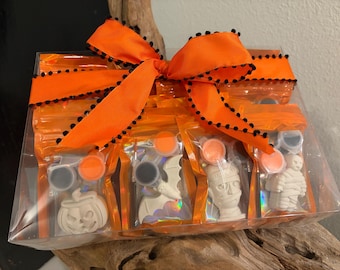 May include: A clear plastic box containing four individual orange foil pouches, each holding a white plaster figure, two small paint pots, and a brush. The figures are a pumpkin, a bat, a mummy head, and a mummy. An orange ribbon with black pom-poms is tied around the box.