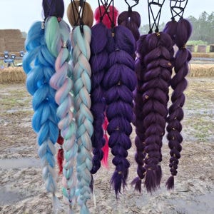 May include: A collection of six synthetic hair extensions styled in thick, chunky braids. The colors include vibrant blue, pastel pink and blue, light blue, and various shades of deep purple. Each braid is secured with small metallic hair cuffs.