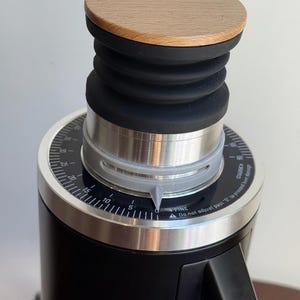 May include: Close-up of a black coffee grinder with a wooden lid and a black rubber bellows. The grinder has a silver adjustment ring with numbers and the text "FINE" and "COARSE".