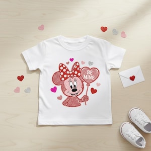 May include: White t-shirt featuring a glittery Minnie Mouse design holding a heart-shaped balloon with the words "BE MINE." The design includes a red polka-dot bow and various heart accents. A white envelope with a heart and white sneakers are also visible.