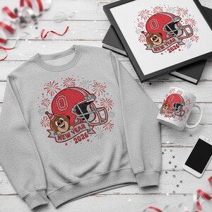 Pode incluir: Um moletom cinza claro, uma impressão emoldurada e uma caneca com um capacete de futebol americano vermelho e um urso de desenho animado usando um chapéu de festa. O design inclui o texto "New Year 2026" e fogos de artifício.