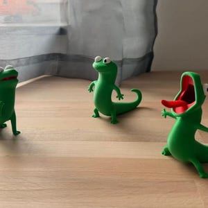 May include: Three green lizard figurines with white eyes and black pupils. The left lizard looks upward, the center lizard stands upright, and the right lizard has an open mouth with a red interior and a protruding red tongue, all in a matte finish.
