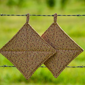 May include: Two square pot holders with a floral pattern in brown and red dots on a tan background hang on a barbed wire fence. The pot holders have a loop for hanging and are set against a blurred green background.