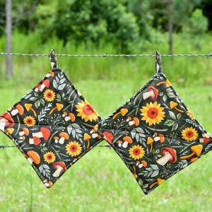 May include: Two square quilted potholders featuring a dark green background with a repeating pattern of orange and white mushrooms, yellow sunflowers, and green foliage. Each potholder has a small fabric loop at the top corner for hanging.